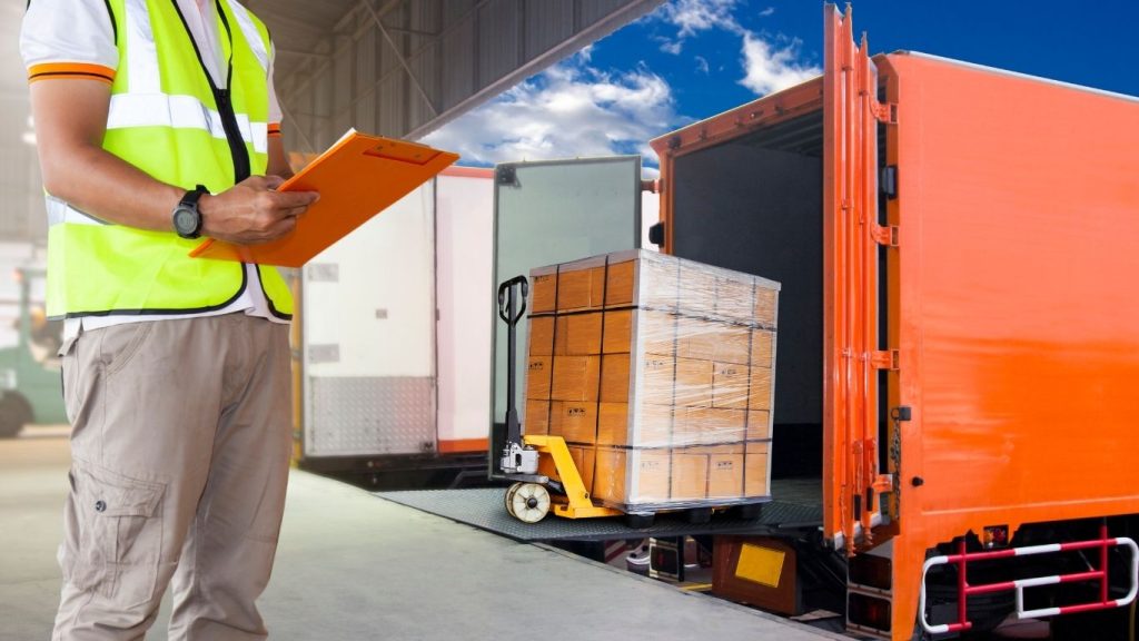 Empresas de frete: caminhão sendo carregado enquanto um homem esta com uma prancheta de conferêrncia