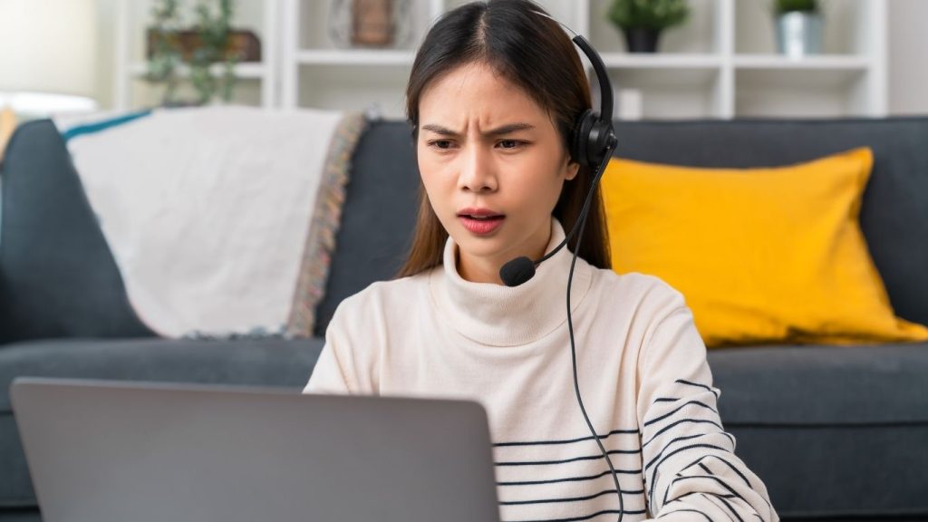 mulher sentada no chão de sua casa enquanto atende uma chamada no fone e olha algo no notebook.